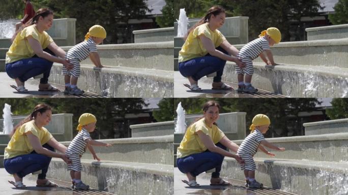 快乐的孩子和他的母亲在阳光明媚的夏日在公园的喷泉里玩得开心。1.5岁的孩子在热天玩喷泉水。