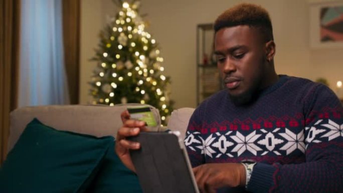 一个非洲外表的年轻人坐在客厅的沙发上，拿着平板电脑和银行卡，在网上商店购物。他仔细输入卡片的数据
