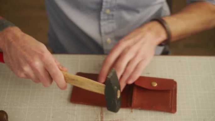皮革工匠在他的车间工作。制作皮革钱包的大师。男人用锤子打孔将持卡人和硬币袋粘在一起。