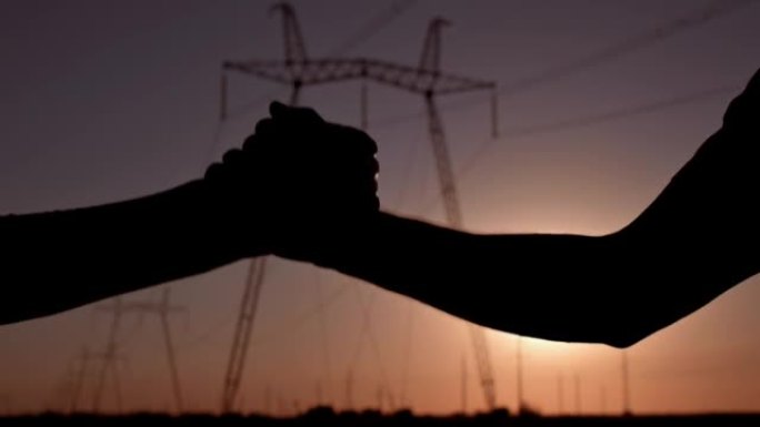 日落时，两名电气工程师在高压电塔的背景下握手。剪影手靠近。团队合作，签订合同，商人之间的协议慢动作