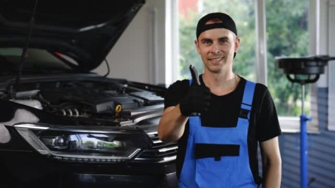 在服务的背景下，汽车车间的汽车修理工肖像显示了竖起大拇指。积极的汽车服务人员对镜头微笑并表现出拇指向