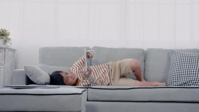4K，慢动作，淘气的亚洲男孩，在客厅里跑着跳上床，以一种有趣的方式，随着伸手拿起平板电脑躺在他附近的