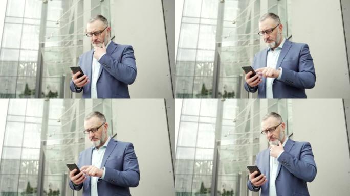 认真体贴的商人站在外面的办公楼附近时使用智能手机。成熟的白发大胡子戴眼镜的高级男性浏览聊天用手机发短
