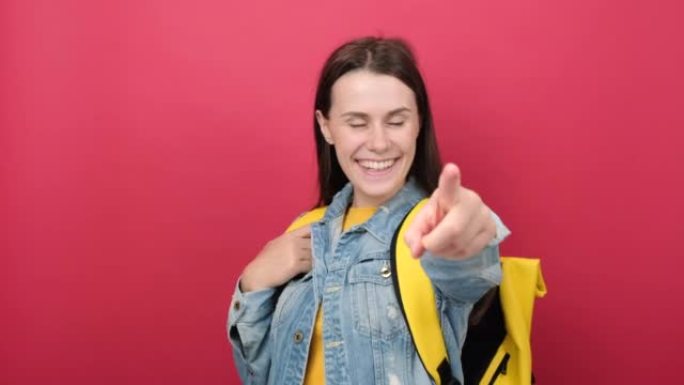 开朗的年轻学生女人穿着牛仔夹克和黄色背包点食指相机在你说做它孤立在工作室的红色背景墙上。高中大学概念