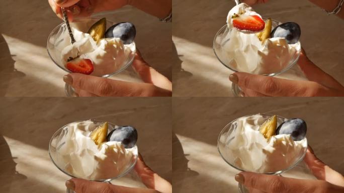 特写女人握着勺子的手吃柔软的奶油冰淇淋，里面有草莓和成熟的李子片。浆果和水果奶油。水果甜点。日落照明