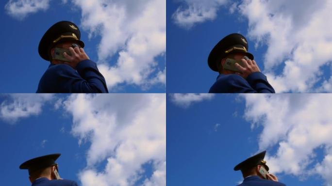 一名戴着帽子的飞行员在机场起飞前正在打电话。飞机的指挥官在蓝天的背景下，手里拿着智能手机与他的亲人交