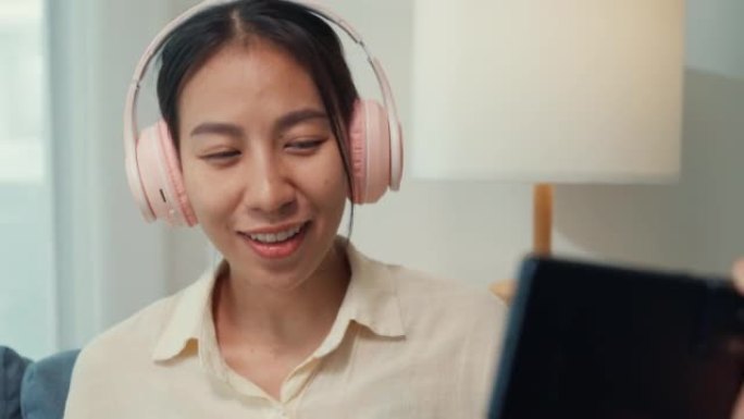 年轻的亚洲女人看沙发上的智能手机戴着耳机听音乐唱歌和欢呼最喜欢的现场乐队音乐会在家里在线音频流广播。