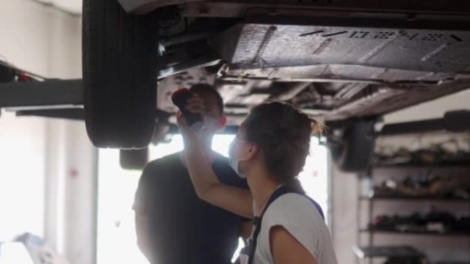 女机械师检查汽车悬架。强壮的女人执教一名男性实习生。工人在升降机上检查车辆的底部和行驶装置。因为检疫