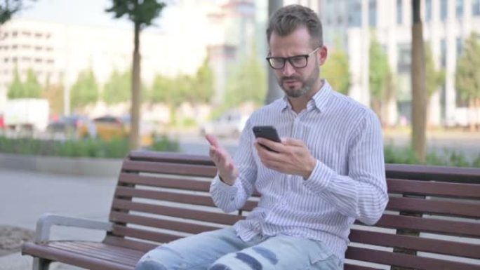年轻的成年男子坐在户外的长凳上玩智能手机