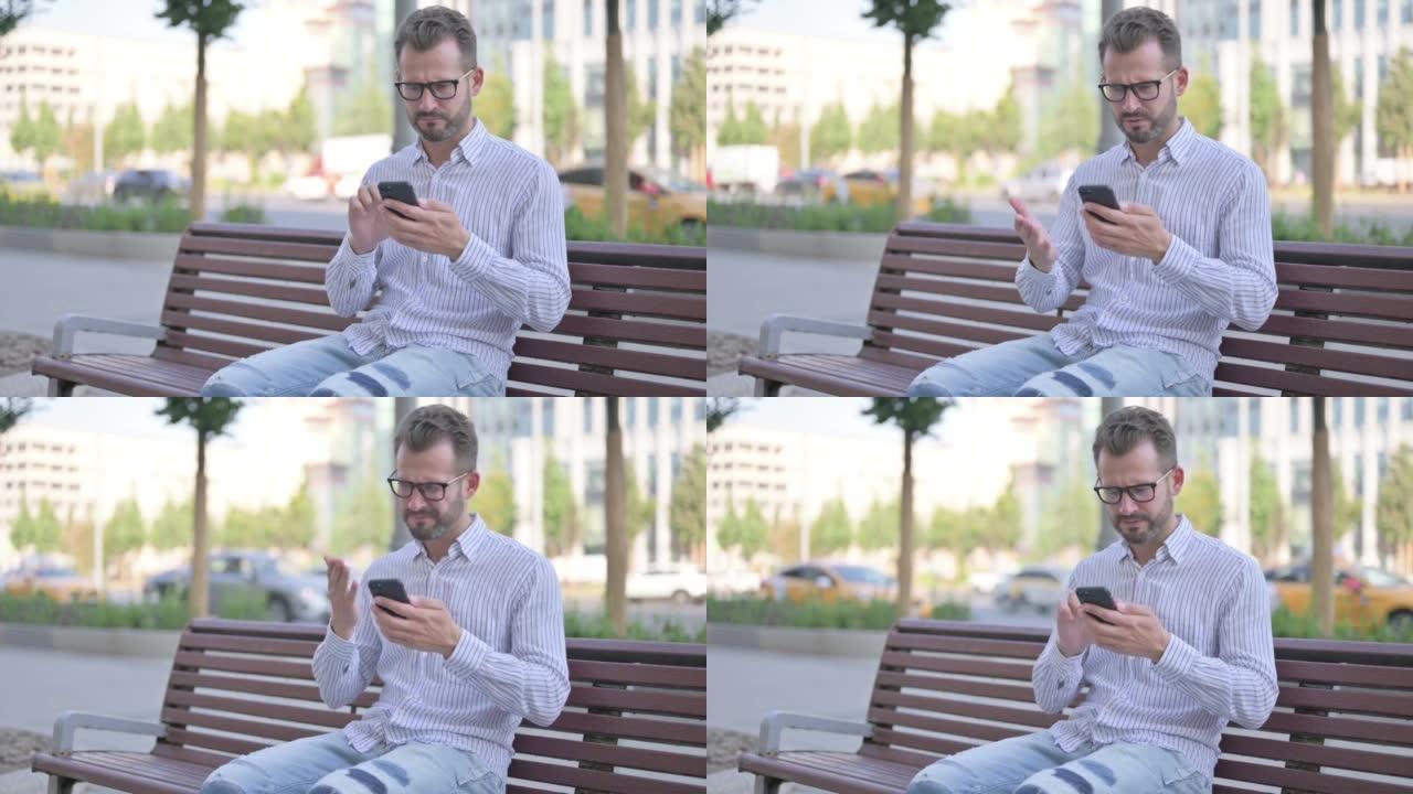 年轻的成年男子坐在户外的长凳上玩智能手机