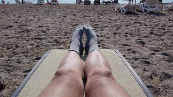 跳舞，扭动袜子中的男性腿和脚趾。一个躺在躺椅上，在海滩上晒日光浴的人的观点。