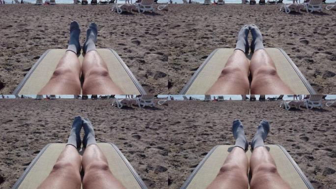 跳舞，扭动袜子中的男性腿和脚趾。一个躺在躺椅上，在海滩上晒日光浴的人的观点。