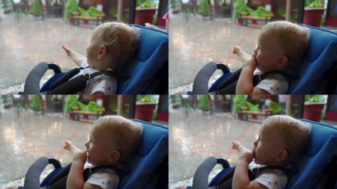 婴儿推车上的孩子看着一场暴雨坐在夏季咖啡馆的树冠下，婴儿的脸顶着雨滴。