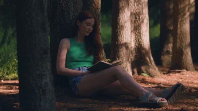 浪漫的年轻女孩看书，坐在户外林地人行道上的树荫下。该国的假期