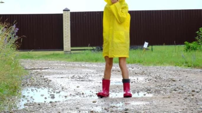 一个穿着红色橡胶靴和黄色雨衣的女孩在村子里下雨后穿过水坑。夏天，自由，童年