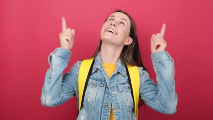 开朗的年轻女学生背着背包，用食指举起伟大的新主意，在工作室的红色背景墙上孤立地摆姿势。高中教育大学学