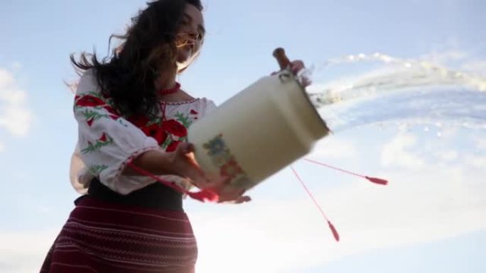 乌克兰妇女穿着传统的民族服装，在天空背景下从罐中倒出水。