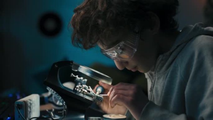 戴着安全眼镜的小男孩，未来的电子技术员，学习如何焊接微处理器电缆和印刷电路，透过放大镜看，修理和测试
