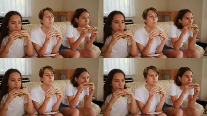 三胞胎姐妹十几岁的女孩在家看电视，坐在沙发上一起吃美味的三明治