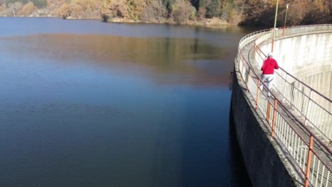 男子在大坝上慢跑，秋天的湖桥，背景是山七彩森林，鸟瞰图