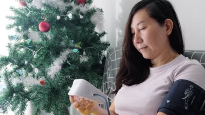 亚洲妇女与血压测试和寻找数字表在网站上的建议，而坐在沙发附近的圣诞树和窗口在家里，健康的生活方式的概
