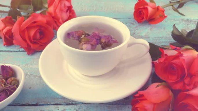 白色的茶帽，带有芬芳的干玫瑰花蕾，新鲜的玫瑰花放在老式的浅蓝色木制桌面上。茶冲泡。