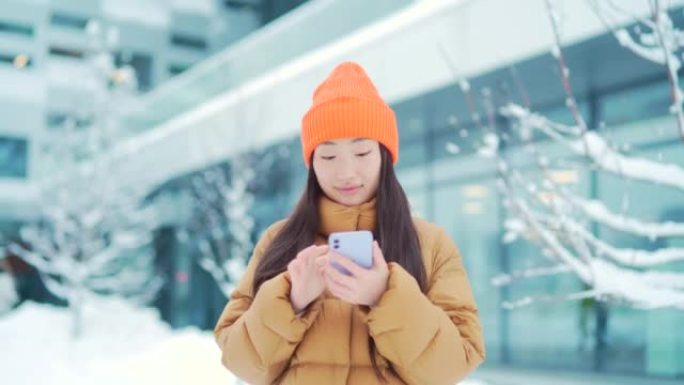 美丽快乐的亚洲青少年女学生站在寒冷的冬季雪城街上使用手机微笑。手里拿着智能手机的年轻女子滚动短信聊天