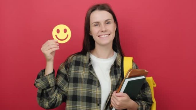 年轻女子学生的肖像拿着小黄纸快乐表情，穿着背包，在工作室里孤立地摆出红色背景，复制空间用于推广内容。