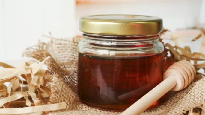 在具有现代厨房背景的木桌上的带有蜂蜜铲斗的玻璃罐中的蜂蜜。慢速平移。