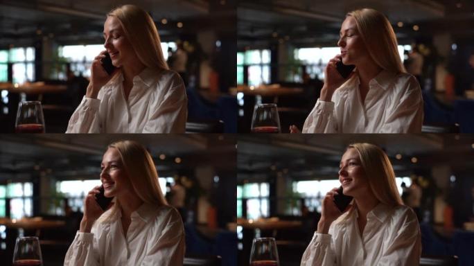 微笑的漂亮金发年轻女子坐在现代餐厅里拿着红酒坐在手机上聊天，室内黝黑。