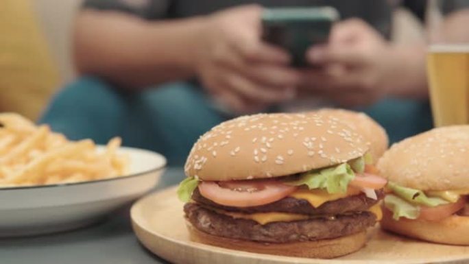 桌子上的汉堡包的4k视频片段，年轻人在家中沙发上使用智能手机，不健康的生活方式概念。