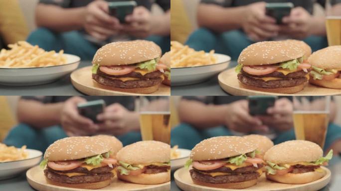 桌子上的汉堡包的4k视频片段，年轻人在家中沙发上使用智能手机，不健康的生活方式概念。