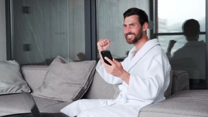 穿着浴袍的兴奋快乐商人在公寓里用电话庆祝胜利，成功赢家盖伊在酒店里看着手机。特写快乐的人在电话上阅读
