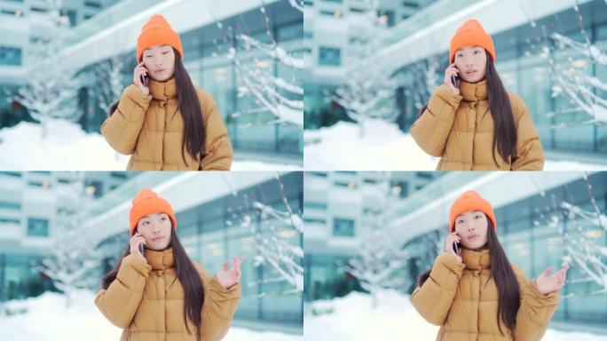 年轻的亚洲妇女在下雪的冬日里用手机说话，校园里的一名学生。快乐女孩对话站在城市街道上的现代城市背景