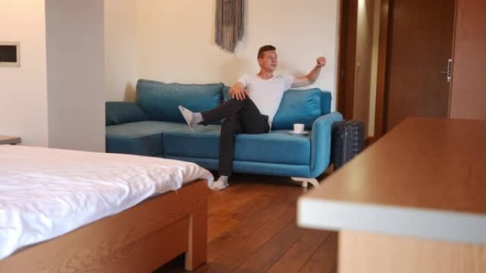 自信的年轻白人男子坐在酒店房间的沙发上，咖啡杯看着远处思考。无忧无虑的英俊游客在室内度假。
