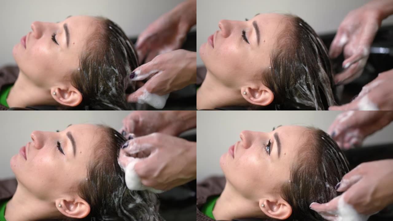 侧视图是发廊里年轻女子的头像，理发师的手在长发上洗发白色泡沫。室内美容院黑发满意的高加索客户肖像。