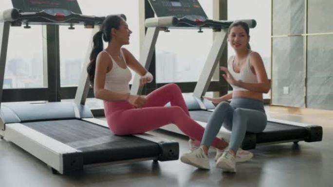 在健身房里跑完步后，在电动跑步机上坐着休息的两位亚洲美女。在健身房里看运动后消耗热量的美女。