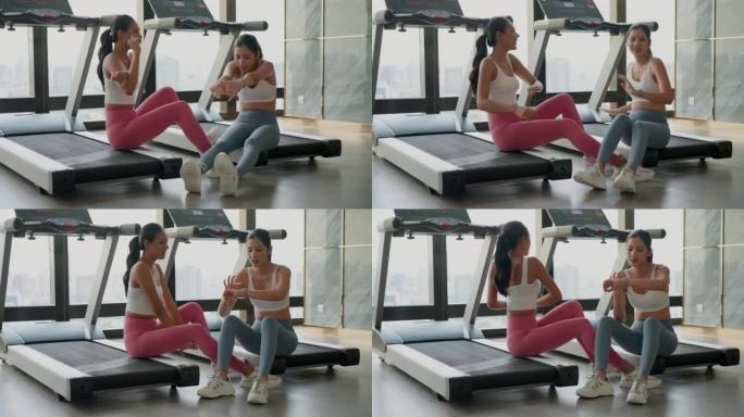 在健身房里跑完步后，在电动跑步机上坐着休息的两位亚洲美女。在健身房里看运动后消耗热量的美女。