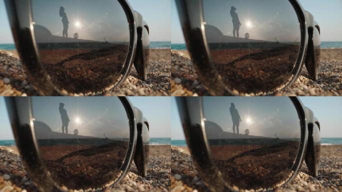 太阳镜躺在大海背景下的沙滩上。女孩在倒影中环顾四周。特写。老瓦探测器。