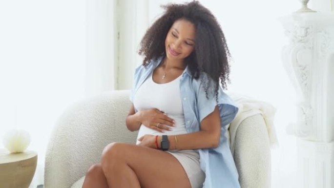 健康怀孕。黑色迷人的孕妇在白色卧室与大窗户坐在椅子上。美国黑人女性妊娠晚期，抚摸腹部。妈妈在等宝贝。