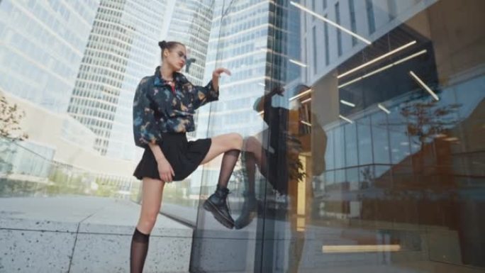 都市风格诱人的女孩穿着彩色风衣短裙和凉爽的未来派眼镜，站在楼梯上，靠腿靠在高层摩天大楼的玻璃墙上。英
