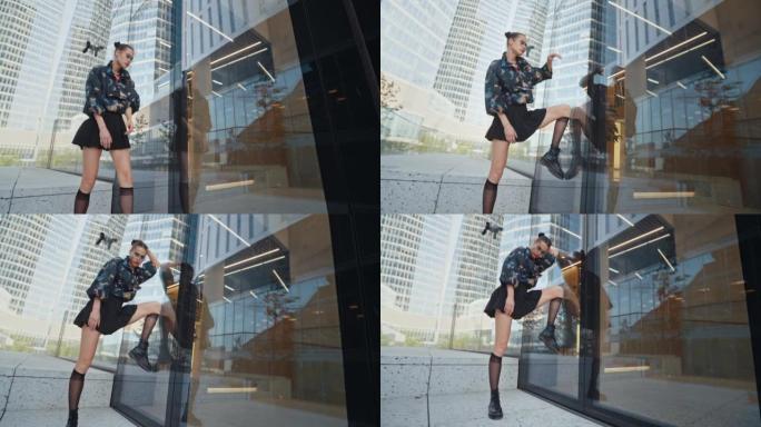都市风格诱人的女孩穿着彩色风衣短裙和凉爽的未来派眼镜，站在楼梯上，靠腿靠在高层摩天大楼的玻璃墙上。英