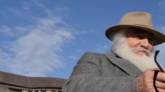 一位白发苍苍的老人，戴着帽子和外套，留着胡须和胡须，手里拿着一本书，坐在公园的长椅上看书。看起来像牧