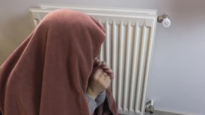 由于能源危机和高汽油价格，女人被毯子覆盖，在散热器旁边变冷。