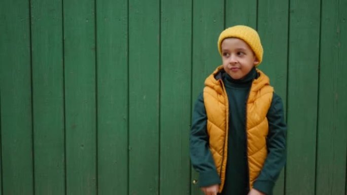 身穿黄色背心，戴着针织帽的男孩孩子在村庄绿色木栅栏的背景下，双手指向左侧。