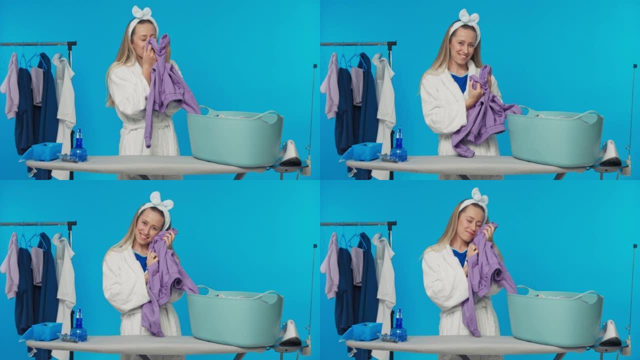 一名妇女在清洁时准备熨烫时，嗅着刚洗干净的闻起来的衣服。工作室蓝色背景上微笑女孩的肖像