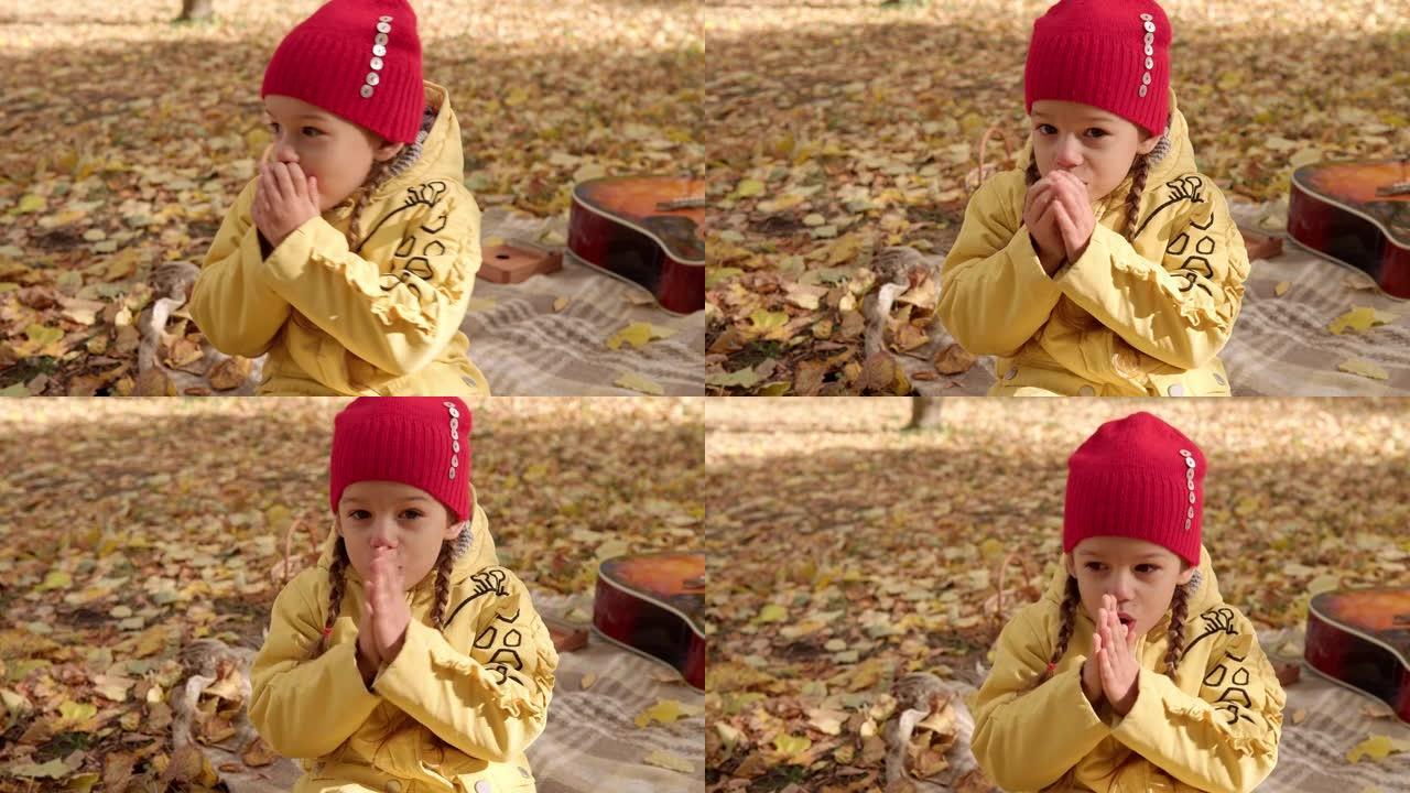 戴着红帽子的可爱的学龄前小女孩温暖着她的手，在篮子里的格子黄色落叶上温暖地吹在她的手掌上呼吸，看着秋