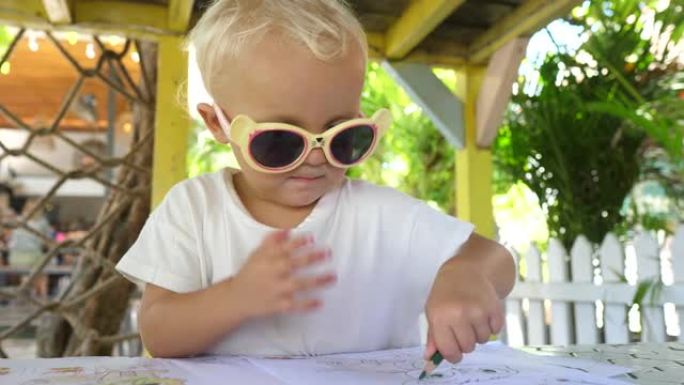 可爱的金发小姑娘，2岁，戴着有趣的黄色太阳镜，用彩色铅笔给彩色书籍着色。儿童游乐场在新鲜空气中进行游