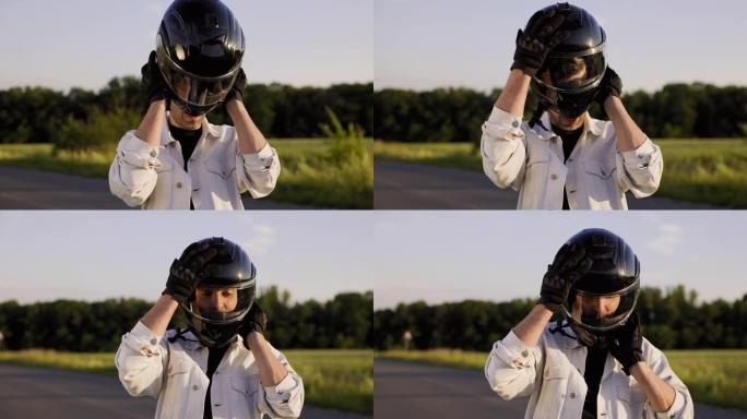 Motorciclist坐在摩托车上，将头盔戴在头上。摩托车站在田野附近的路上。日落背景