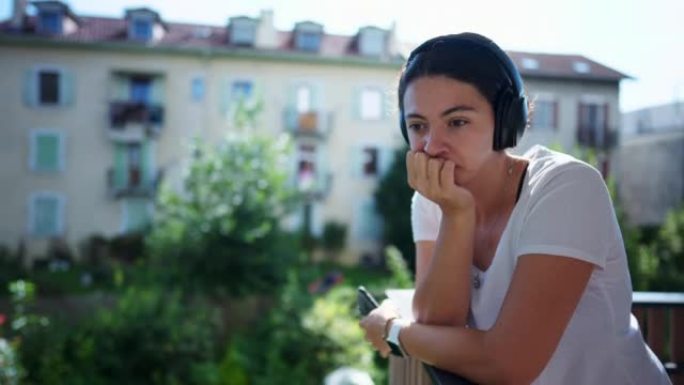 女人戴着耳机站在公寓阳台外沉思。听播客有声读物或音乐的人。带降噪耳机的成年女孩